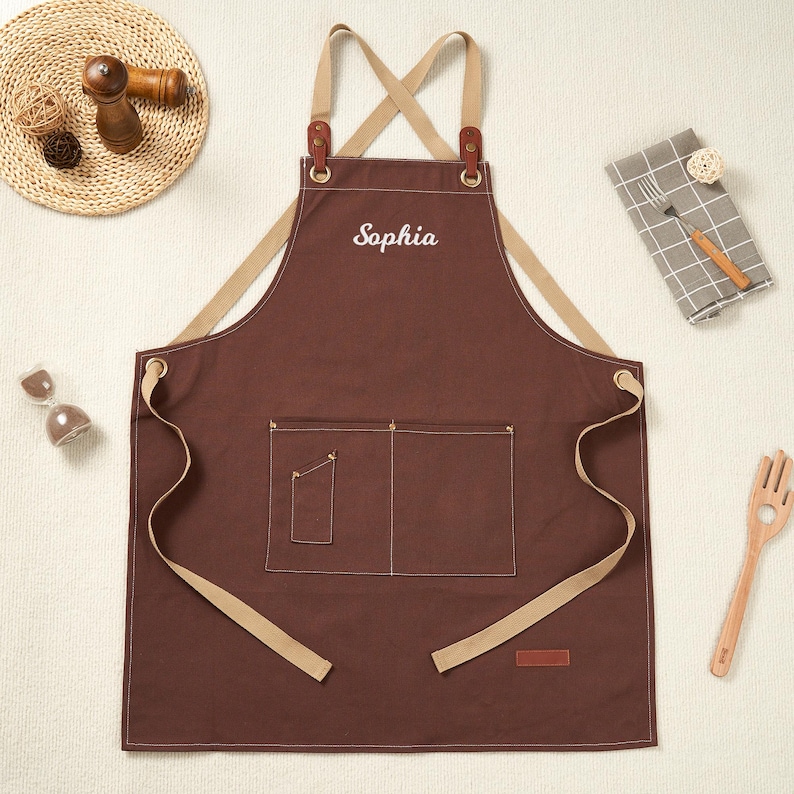 May include: A brown apron embroidered with the name "Sophia" in white. It has two front pockets, tan straps, and leather details. Ideal for cooking or crafting, the apron is a practical accessory.