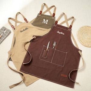 May include: Three personalised aprons in tan, olive green, and brown. Each apron has a different name embroidered on it: "Isabella", "Matthew", and "Sophia". The aprons have cross-back straps with leather accents and front pockets. A knife and fork are in the front pocket of the brown apron.