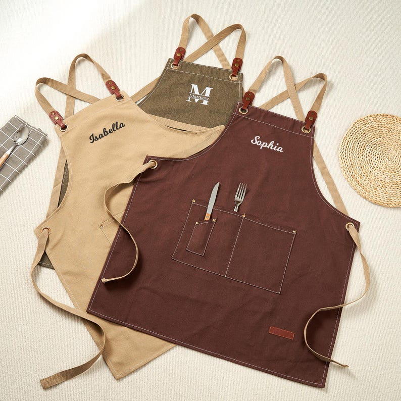 May include: Three personalised aprons in tan, olive green, and brown. Each apron has a different name embroidered on it: "Isabella", "Matthew", and "Sophia". The aprons have cross-back straps with leather accents and front pockets. A knife and fork are in the front pocket of the brown apron.