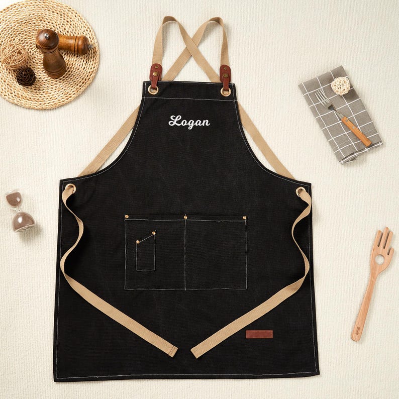 May include: Black apron with tan straps and pockets. The apron has the name "Logan" embroidered in white. The apron has a leather patch and metal grommets. The apron is designed for cooking and crafting.