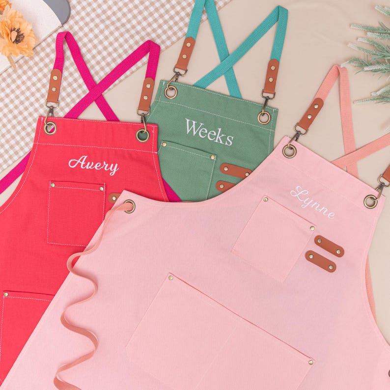 May include: Three personalized aprons in red, green, and pink. Each apron has a front pocket, adjustable straps, and the names "Avery", "Weeks", and "Lynne" embroidered on them. The straps are a contrasting color and have brown leather accents.