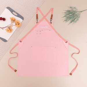 May include: A light pink apron with a cross-back design, featuring brown leather accents and a front pocket. The apron has long straps and a small embroidered detail. The apron is designed for kitchen or craft use.