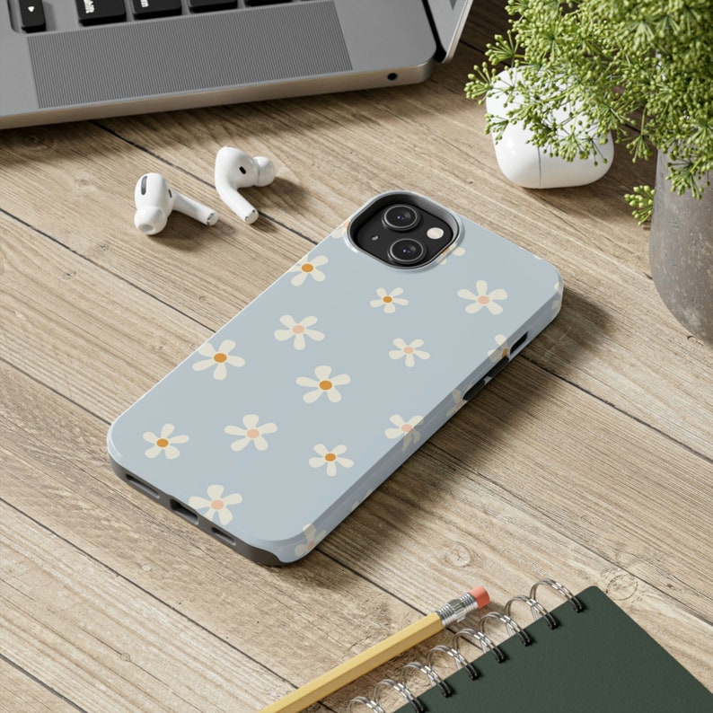 Puede incluir: Una funda de tel&eacute;fono azul claro con un patr&oacute;n de margaritas blancas. La funda tiene un recorte para el objetivo de la c&aacute;mara.