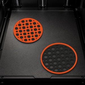 May include: Two circular coasters with a geometric star-patterned cutout design. One coaster features an orange frame with a black interior, while the other has a black frame with an orange interior, both resting on a flat black surface.