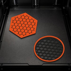 Op de afbeelding: Een oranje zeshoekige onderzetter met een zwart honingraatpatroon en een ronde onderzetter met een zwart honingraatcentrum en een oranje rand, beide rustend op een plat zwart oppervlak in een 3D-printer.