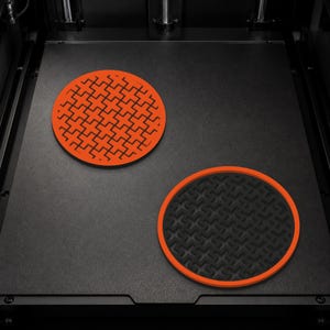 Op de afbeelding: Twee ronde onderzetters op een plat oppervlak. De ene onderzetter is feloranje met een uitgesneden geometrisch kruispatroon. De tweede onderzetter is zwart met een getextureerd oppervlak en een effen feloranje cirkelvormige rand.