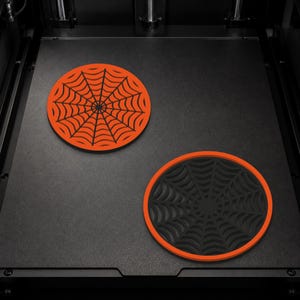 Könnte beinhalten: Zwei kreisförmige Untersetzer mit Spinnennetz-Motiv. Ein Untersetzer ist orange mit schwarzen Netzlinien, der andere ist schwarz mit einem orangefarbenen Rand und schwarzen Netzlinien. Sie liegen auf einer flachen, dunklen, strukturierten Fläche.