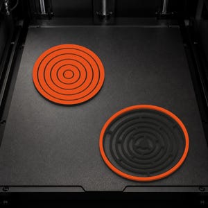 May include: Two circular objects resting on a flat black surface. One object features a bright orange base with concentric black rings. The second object has a bright orange outer ring surrounding a complex black circular maze pattern.
