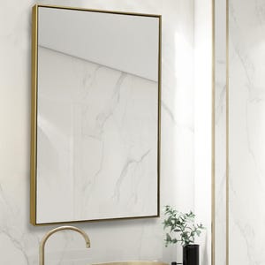 May include: A gold-toned oval vessel sink with a brushed metallic finish, paired with a tall, curved gold faucet. The sink sits on a white marble countertop featuring grey veining, positioned below a rectangular mirror with a thin gold frame.