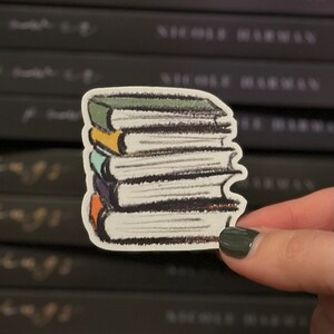 Puede incluir: Una pegatina de una pila de libros con una encuadernación verde, amarilla, azul y naranja. La pegatina está sobre un fondo de una estantería con libros negros.
