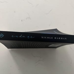 May include: A black book titled "endure" by Nicole Harman. The title is in a teal script font, and the author's name is in a white sans-serif font. The book has a teal diamond-shaped logo.