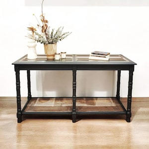 May include: A black wooden console table with a glass top and a lower shelf with woven cane. The table has four legs and is decorated with a vase of dried flowers and candles.