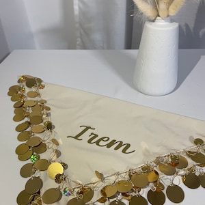 May include: Cream-coloured scarf with gold sequin trim and the name "Irem" printed in gold. A white vase with dried flowers is in the background. The scarf is draped on a white surface.