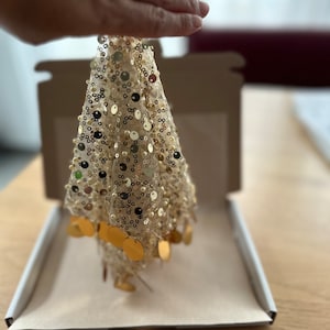 Puede incluir: Un pequeño árbol de Navidad decorativo hecho de malla dorada y adornado con lentejuelas y adornos dorados en forma de disco. El árbol está sostenido en la mano de una persona y está dentro de una caja de cartón.