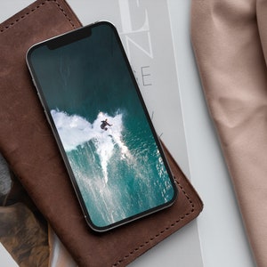 May include: A brown leather wallet with a black smartphone on top. The phone screen shows a photo of a person surfing a wave.