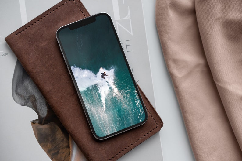 May include: A brown leather wallet with a black smartphone on top. The phone screen shows a photo of a person surfing a wave.