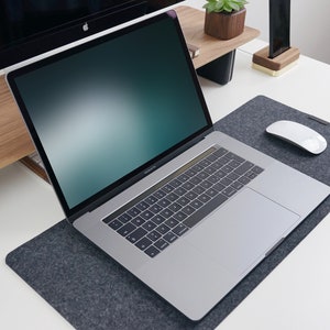 May include: A silver laptop computer with a black keyboard and a gray trackpad is on a gray felt desk mat. A white wireless mouse is on the desk mat. The laptop is open and the screen is displaying a blurred green and blue background.