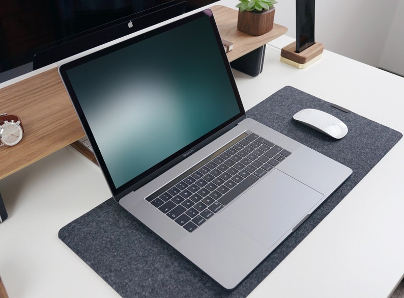 May include: A silver laptop computer with a black keyboard and a gray trackpad is on a gray felt desk mat. A white wireless mouse is on the desk mat. The laptop is open and the screen is displaying a blurred green and blue background.