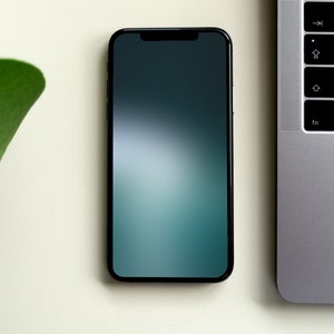 May include: A black smartphone with a dark green and blue gradient screen is lying on a white surface next to a silver laptop.