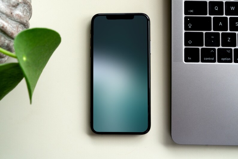 May include: A black smartphone with a dark green and blue gradient screen is lying on a white surface next to a silver laptop.