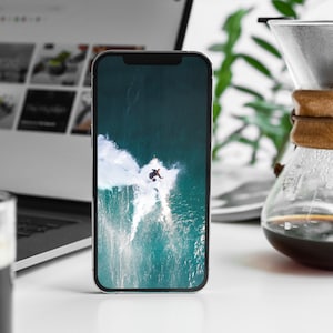 May include: A white smartphone with a black screen displaying a photo of a person surfing a large wave. The wave is a turquoise blue color with white foam.