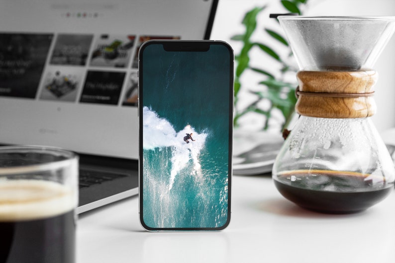 May include: A white smartphone with a black screen displaying a photo of a person surfing a large wave. The wave is a turquoise blue color with white foam.