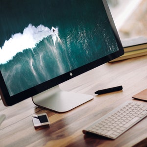 May include: A silver desktop computer with a black screen displaying a photo of a surfer riding a large wave. The computer is on a wooden desk with a white keyboard, a brown leather mouse pad, and a white mouse.