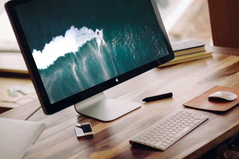 May include: A silver desktop computer with a black screen displaying a photo of a surfer riding a large wave. The computer is on a wooden desk with a white keyboard, a brown leather mouse pad, and a white mouse.