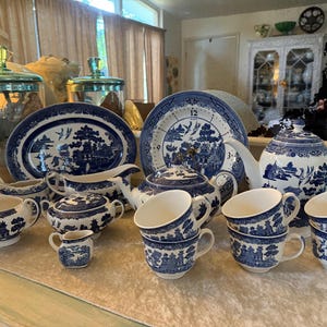 May include: A collection of blue and white porcelain tableware, including a teapot, cups, saucers, a serving platter, and a sugar bowl. The pieces feature a classic Willow pattern design. The set is arranged on a light-colored surface.