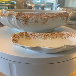 May include: A white and brown floral patterned serving bowl with handles and a matching small dish. The bowl is oval-shaped and sits on a white surface. The dish is also oval-shaped with a gold trim.