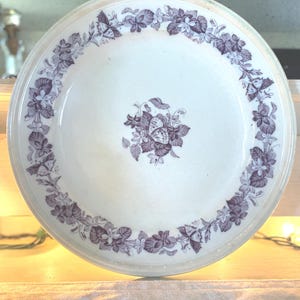 May include: A round white ceramic plate featuring a dark purple transferware pattern. The rim is decorated with a continuous floral and butterfly motif, while the center displays a cluster of leaves, small flowers, and a single butterfly.