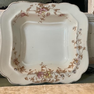 May include: Cream-colored, square antique dish with a scalloped edge. The dish features floral patterns in pink, yellow, and brown. The design is symmetrical, with floral arrangements in each corner and along the sides.