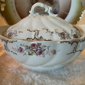 May include: A white porcelain serving bowl with a lid, featuring a floral design in pink, purple, and yellow. The bowl has a scalloped edge and a decorative handle on the lid. The rim and handle are accented with gold.