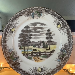May include: A white ceramic plate with a brown border featuring a pastoral scene. The center depicts a house, trees, and a fence. The border has floral and grape designs. A decorative piece for serving or display.