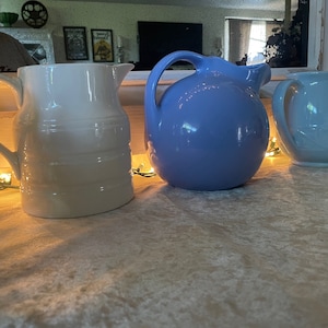May include: Three ceramic pitchers in varying shades of white and blue are arranged on a table. The white pitcher has a ribbed design, the blue pitcher is a solid color, and the light blue pitcher has a floral design.