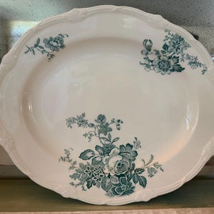 May include: An oval white ceramic serving platter featuring a scalloped edge and a decorative teal floral pattern. The design includes a large cluster of flowers at the bottom and smaller floral sprigs positioned on the upper left and right sides.