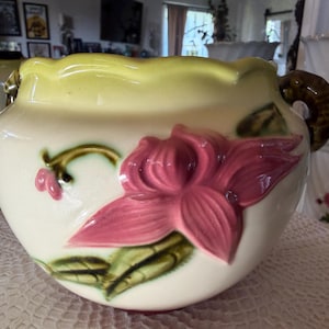 May include: Cream-colored ceramic planter with a large pink flower and green leaves. The rim is light yellow, and it has brown handles. A smaller planter is visible in the background.