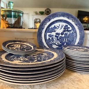 May include: A set of blue and white ceramic plates featuring the Willow pattern. The image shows a stack of dinner plates, side plates, and smaller plates. The plates are displayed on a light-coloured surface.