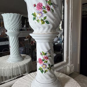 May include: White ceramic planter with a pedestal base. The planter features a spiral design and is decorated with pink, purple, and yellow flowers and a butterfly. The rim and base have gold trim. A second planter is visible in the background.