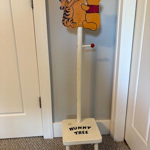 May include: A vintage wooden coat rack featuring Winnie the Pooh and Tigger. The rack has a small stool base with "HUNNY TREE" written on it. The top has a red balloon with the word "POOH" on it.