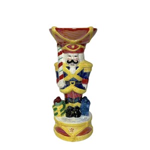 May include: A ceramic nutcracker candle holder with a red, yellow, blue, and white paint scheme. The nutcracker is holding a candy cane and has a red hat. The base of the holder is decorated with a star and a red and yellow design.