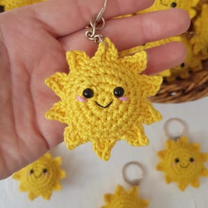 May include: Yellow crocheted sun keychains with smiling faces. The keychains are handmade and have black eyes and pink cheeks. A basket filled with similar keychains is in the background.