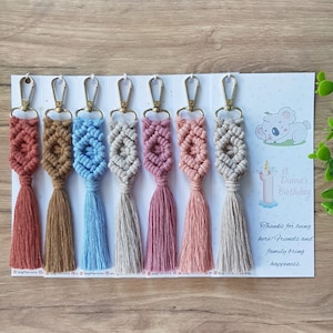Könnte beinhalten: Sieben bunte Makramee-Schlüsselanhänger mit Quasten, jeder mit einer anderen Farbe und einem anderen Muster. Die Schlüsselanhänger werden auf einer weißen Karte mit einer Koala-Illustration und dem Text "Es ist Dianas 1. Geburtstag" und "Danke, dass ihr da seid! Freunde und Familie bringen Glück." angezeigt.
