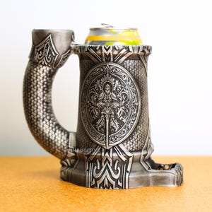 May include: A silver-coloured, decorative beer stein with an ornate handle and base. The stein features a detailed embossed design of a warrior with a sword. A can of beverage sits inside the stein. The stein has a small dice tray.