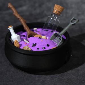 May include: A black cauldron filled with purple sand, containing a brown twig, a small white bottle, a clear glass vial with a cork stopper, and a grey plastic shovel. The purple sand is decorated with small black spider shapes and tiny red beads.