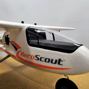 May include: AeroScout model airplane, white with black and orange accents. The plane has a clear cockpit and black propeller. The text "AeroScout" is printed on the side of the plane.