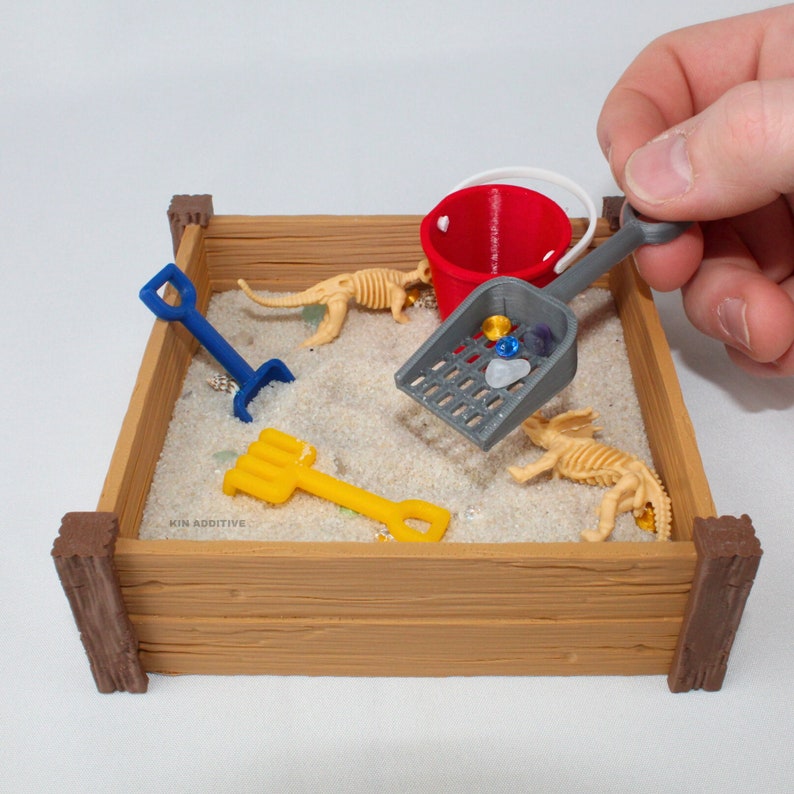 May include: A miniature wooden sandbox with a red bucket, a gray scoop, a yellow rake, a blue shovel, and two white plastic dinosaur skeletons. The sandbox has the text "KIN ADDITIVE" printed on the side.