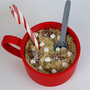 May include: A red mug filled with tan sand, marshmallows, and gingerbread men. A candy cane and gray spoon are also in the mug. The mug is on a white surface. Festive holiday theme.