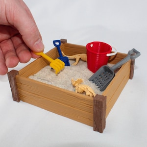 May include: A wooden sandbox with a red bucket, a blue shovel, a yellow rake, and two plastic dinosaur skeletons. The sandbox is made of wood and has a brown stain.