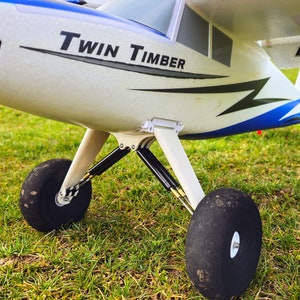 以下が含まれることがあります： 白と青の双発機で、側面に「Twin Timber」と印刷されています。飛行機は芝生のフィールドにあり、黒い車輪が付いています。